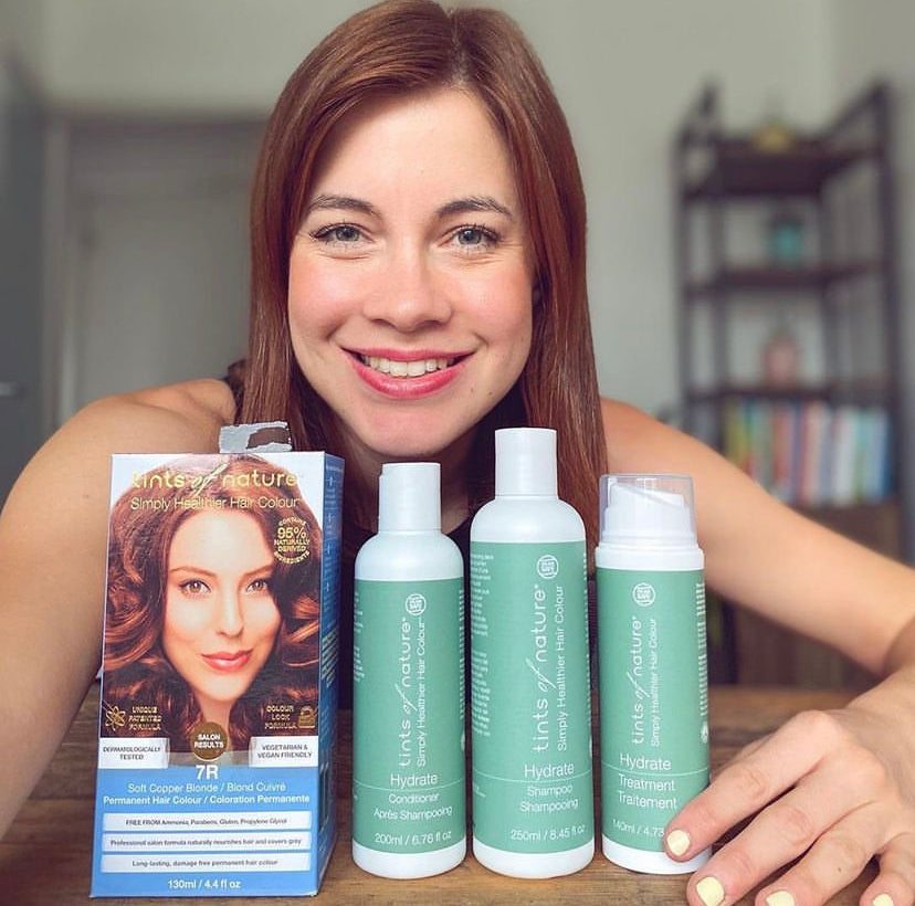 A woman smiles at the camera while displaying vegan haircare products, including a box of Tints of Nature hair dye and Hydrate shampoo, conditioner and treatment, on a table in front of her.