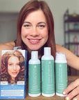 A woman smiles at the camera while displaying vegan haircare products, including a box of Tints of Nature hair dye and Hydrate shampoo, conditioner and treatment, on a table in front of her.