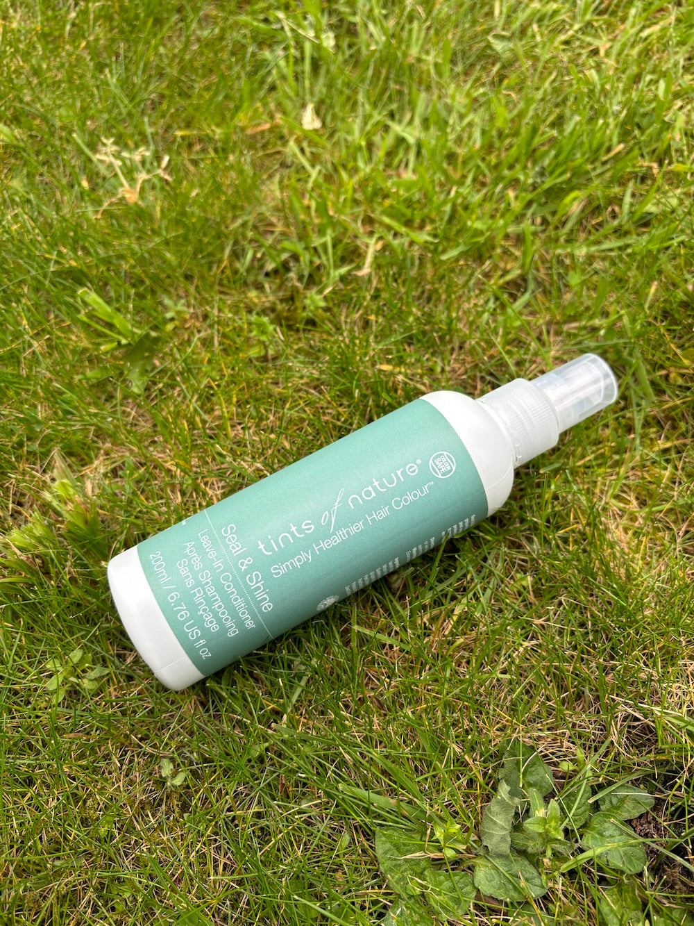 Tints of Nature Seal and Shine Leave-in Conditioner spray bottle resting on green grass outdoors.