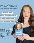 Happy woman holding Tints of Nature permanent hair dye carton with text on screen; covers greys, vibrant long-lasting results, salon-quality, 100% satisfaction guarantee